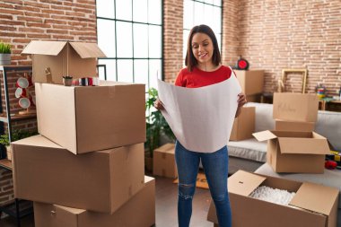 Young beautiful hispanic woman smiling confident reading house plans at new home