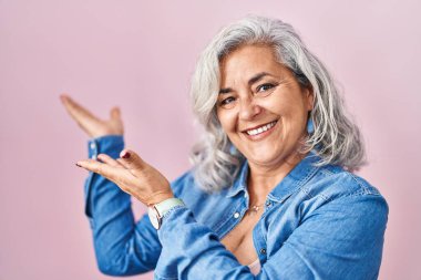 Middle age woman with grey hair standing over pink background inviting to enter smiling natural with open hand 