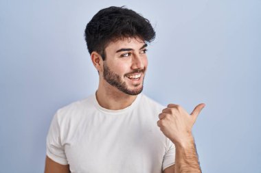 Hispanic man with beard standing over white background smiling with happy face looking and pointing to the side with thumb up. 
