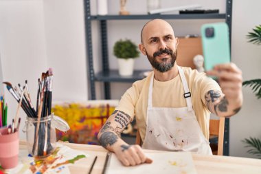 Genç kel adam gülümsüyor. Sanat stüdyosundaki akıllı telefondan özçekim yapıyor.