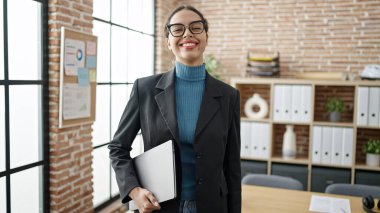 Young beautiful hispanic woman business worker smiling confident holding laptop at office