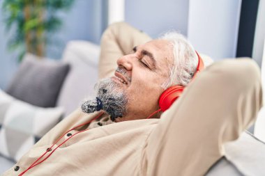 Orta yaşlı, kır saçlı, müzik dinleyen adam evdeki kanepede oturuyor.