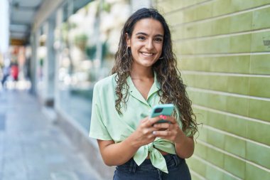 Genç, güzel, İspanyol bir kadın sokakta akıllı telefon kullanarak gülümsüyor.