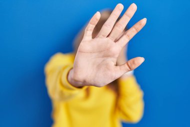 Mavi arka plan üzerinde duran genç beyaz kadın elleriyle gözlerini kapatıyor ve üzgün ve korku ifadeleriyle jestleri durduruyor. utangaç ve olumsuz kavram. 