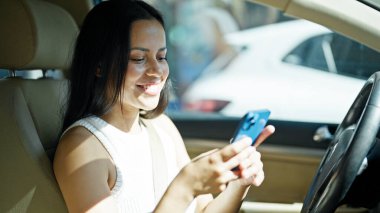 Genç, güzel, İspanyol bir kadın akıllı telefon kullanıyor. Sokakta arabada oturuyor.