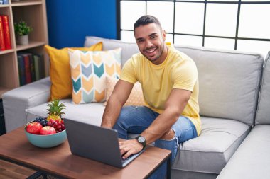 Genç İspanyol adam evdeki koltukta dizüstü bilgisayar kullanıyor.