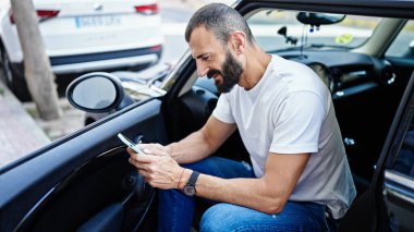 Young hispanic man using smartphone sitting on car at street