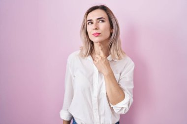 Young beautiful woman standing over pink background thinking concentrated about doubt with finger on chin and looking up wondering 