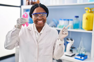 Afrika kökenli Amerikalı bir kadın, elinde pembe kurdeleyle laboratuvarda çalışıyor. 