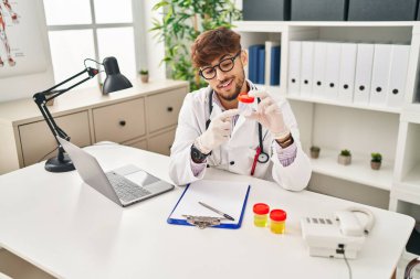 Young arab man wearing doctor uniform analysing urine test tube speaking at clinic