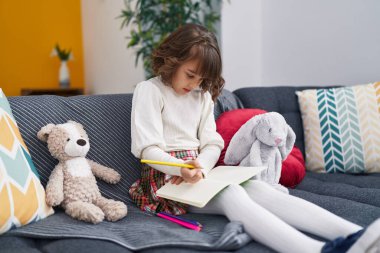 Adorable hispanic girl drawing on notebook sitting on sofa at home