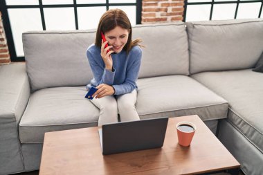 Akıllı telefondan konuşan genç sarışın kadın elinde kredi kartıyla evdeki kanepede oturuyor.