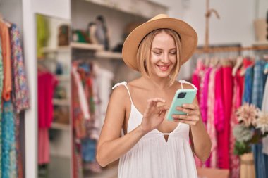 Young blonde woman customer wearing summer hat using smartphone at clothing store