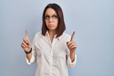 Young hispanic woman standing over white background pointing up looking sad and upset, indicating direction with fingers, unhappy and depressed. 