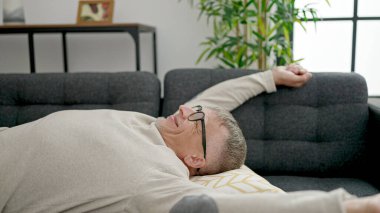 Orta yaşlı, kır saçlı adam evdeki kanepede dinleniyor.