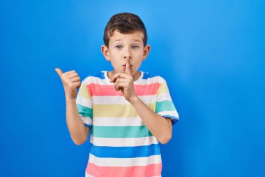 Mavi arka planda dikilen beyaz bir genç sessiz olmak istiyor. Parmakları dudaklarında, elleri yandan işaret ederek. sessizlik ve gizli konsept. 