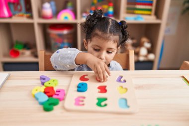 Adorable hispanic girl playing with maths puzzle game sitting on table at kindergarten