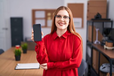 Ofiste çalışan kızıl saçlı kadın akıllı telefon ekranı gösteriyor. Pozitif ve mutlu görünüyor. Kendine güvenen bir gülümsemeyle dişlerini gösteriyor. 