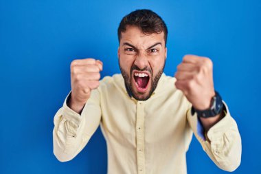 Handsome hispanic man standing over blue background angry and mad raising fists frustrated and furious while shouting with anger. rage and aggressive concept.  clipart