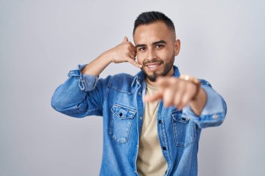 Young hispanic man standing over isolated background smiling doing talking on the telephone gesture and pointing to you. call me.  clipart