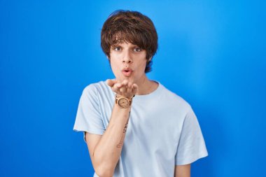 Latin asıllı genç bir adam mavi arka planda durup kameraya bakarak havada bir öpücük savuruyor ve sevimli ve seksi oluyor. aşk ifadesi. 