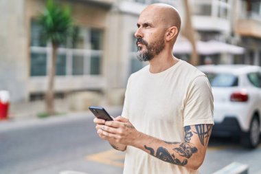 Genç kel adam akıllı telefon kullanıyor ve sokakta ciddi bir yüz ifadesi var.