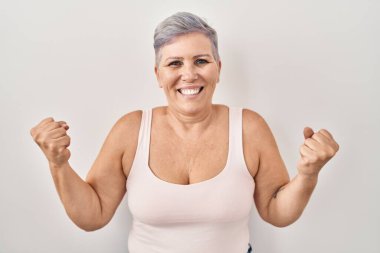 Beyaz arka planda duran orta yaşlı beyaz bir kadın çok mutlu ve heyecanlı bir şekilde kollarını kaldırıp, gülümseyerek ve başarı için bağırarak kazanan jestler yapıyor. kutlama konsepti. 