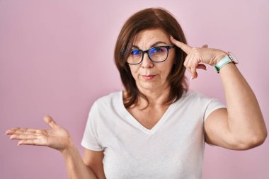 Middle age hispanic woman standing over pink background confused and annoyed with open palm showing copy space and pointing finger to forehead. think about it. 