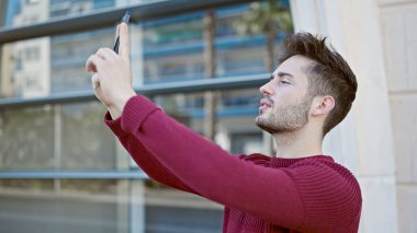 Genç İspanyol adam caddedeki akıllı telefonun yanında fotoğraf çekiyor.