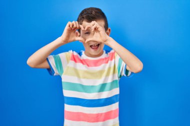 Mavi arka planda duran, elleri ve parmaklarıyla kalp şeklinde gülümseyen beyaz bir çocuk. 