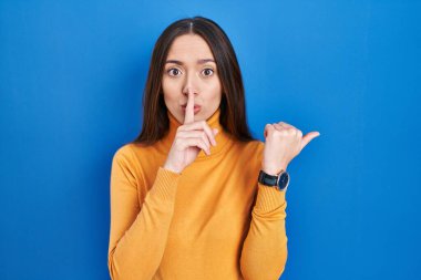 Genç esmer kadın mavi arka planda dikiliyor ve sessiz olmak istiyor. Parmakları dudaklarında, elleri yandan işaret ederek. sessizlik ve gizli konsept. 