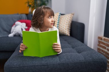 Adorable hispanic girl reading book lying on sofa at home