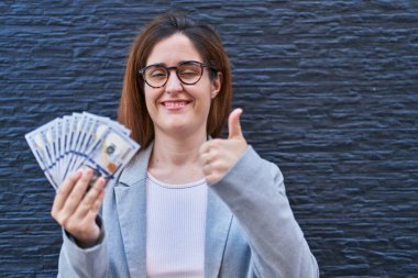 Elinde dolarlar tutan esmer kadın mutlu ve pozitif gülümsüyor, başparmağıyla mükemmel ve onay işareti yapıyor. 