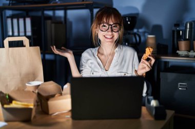 Genç ve güzel bir kadın bilgisayar bilgisayarı kullanıyor ve yemek yiyor. Gülümsüyor, gülümsüyor ve avuç içi kameraya bakıyor.. 