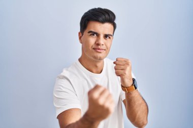 Hispanic man standing over blue background ready to fight with fist defense gesture, angry and upset face, afraid of problem 