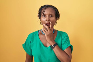 Sarı arka planda duran Afrikalı kadın, elleri çenesinde, inanamama, şaşkınlık ve şaşkınlık dolu bir ifade takınıyor. 