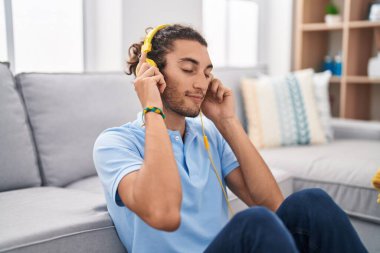 Young hispanic man listening to music sitting on floor at home