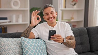 Young hispanic man using smartphone doing ok gesture at home