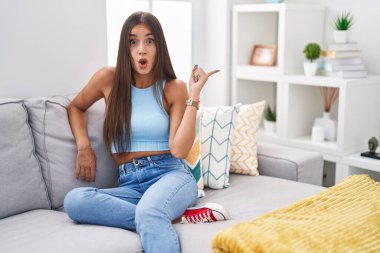 Evdeki kanepede oturan genç esmer kadın şaşkın şaşkın, ağzı açık, parmaklarını kenarı işaret ederek şaşırdı.. 