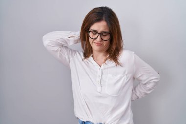 Esmer kadın beyaz arka planda duruyor. Boyun ağrısı çekiyor, boynuna elle dokunuyor, kas ağrısı çekiyor. 