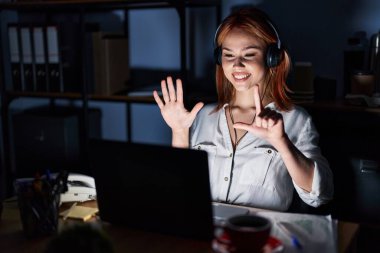 Young caucasian woman working at the office at night showing and pointing up with fingers number seven while smiling confident and happy. 