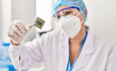 Orta yaşlı kadın, laboratuarda Cpu işlemcisi tutan bilim adamı üniforması ve tıbbi maske takıyor.