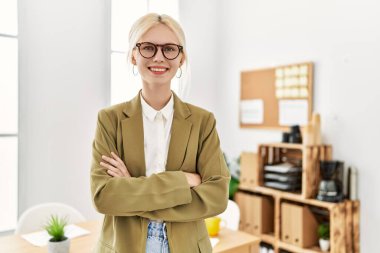 Genç, sarışın, iş kadını, gülümseyen, kendine güvenen, kollarını kavuşturup ofiste el kol kola duran.