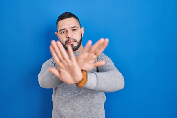 青の背景の拒絶表現の上に立つヒスパニック系の男腕と手のひらを横断負の記号を行う 怒っている顔 — ストック写真