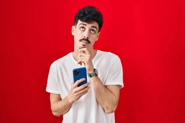 西班牙裔男人在红色背景上用智能手机 手托着下巴思考问题 沉思的表情 脸上挂着沉思的笑容 怀疑概念 — 图库照片