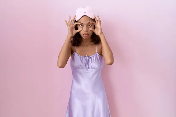 Young hispanic woman wearing sleep mask and nightgown trying to open eyes with fingers, sleepy and tired for morning fatigue