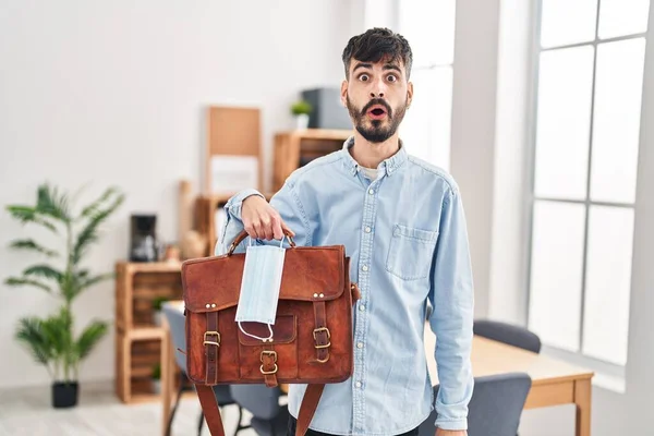 Stock image Young hispanic man with beard working at the office holding suitcase and safety mask scared and amazed with open mouth for surprise, disbelief face 