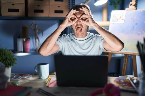 中年男子坐在艺术工作室里 晚上拿着笔记本电脑 做着像双筒望远镜一样的手势 伸出舌头 眼睛透过手指看着 疯狂的表达 — 图库照片