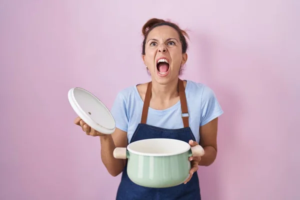 Μπρουνέτ Γυναίκα Φορώντας Ποδιά Κρατώντας Κατσαρόλα Μαγειρέματος Θυμωμένος Και Τρελός — Φωτογραφία Αρχείου
