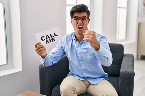 Hispanic man working at therapy office holding call me banner annoyed and frustrated shouting with anger, yelling crazy with anger and hand raised